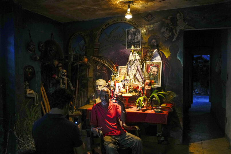 FILE - Juan Gonzalez sits next to his altar adorned Santeria and Catholic deities along with a photo of the late Cuban leader Fidel Castro, at his home in El Cobre, Cuba, Feb. 10, 2024. Diverse beliefs can be found mixed together on altars in Cuban homes, with the Virgin Mary sharing space with a ceramic Buddha and a warrior spirit from the Afro-Cuban faith. (AP Photo/Ramon Espinosa, File)